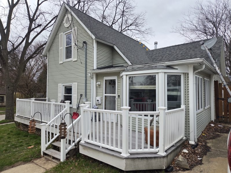 Aerial view of a brick house with a dark shingle roof showing chalk circles marking hail damage, surrounded by green trees and lawn.